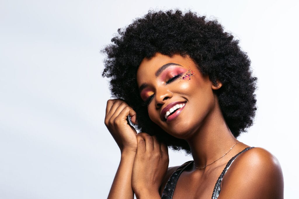 Smiling woman with natural curly hair showcasing flawless skin and elegant makeup, posing with one hand near her face.