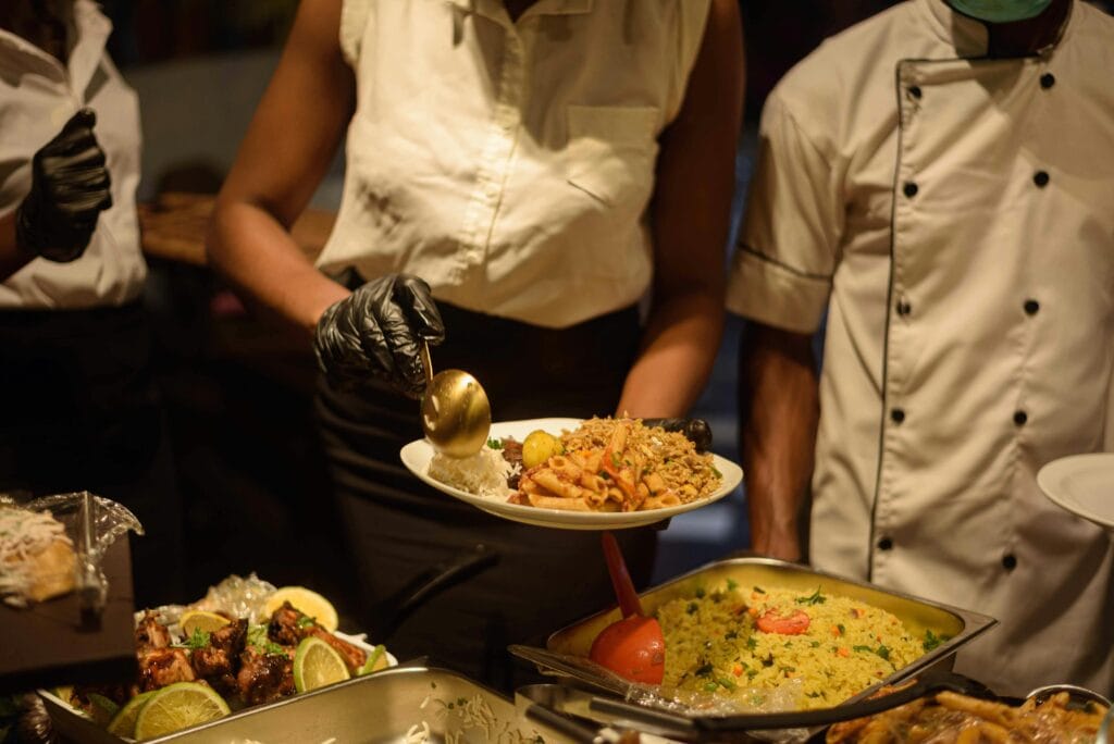 A caterer serves a plate of assorted traditional foods, including rice, pasta, and vegetables, at a buffet during a formal event.