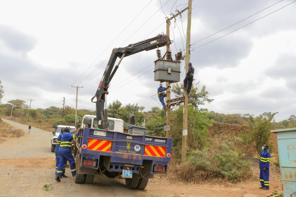 Technicians from Kenya Power installing or maintaining an electrical transformer on a rural utility pole using a service truck with a crane.